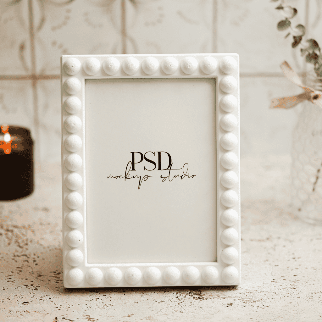 A white, beaded photo frame stands on a rustic surface next to a lit candle in a dark glass holder and a vase with greenery. The frame displays a mockup studio logo on a blank background.