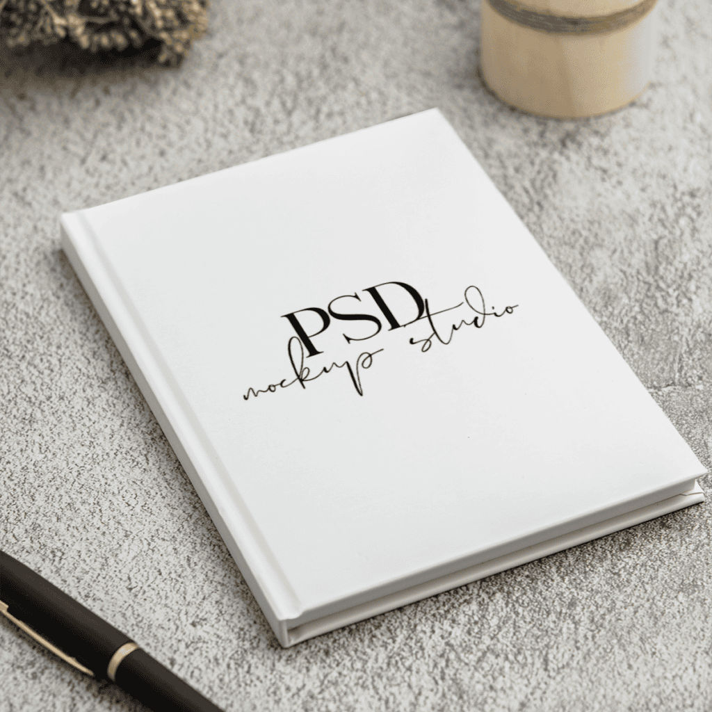 A white hardcover book with “PSD mockup studio” written on the cover rests on a textured gray surface, near a black and gold pen and a round wooden object.