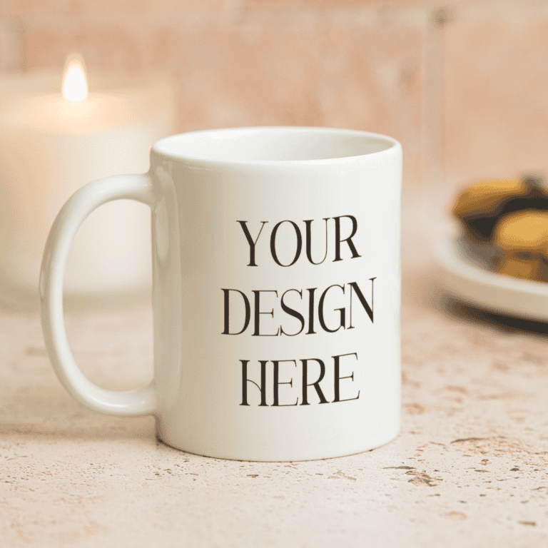 A white ceramic mug with the words YOUR DESIGN HERE printed on it sits on a light textured surface. In the background, there is a lit candle and a plate of cookies, both softly out of focus.