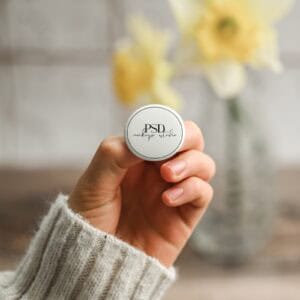 A hand in a cream knit sweater holds a small round container labeled PSD makeup studio, resembling the "1.25 Inch Button Pin Mockup, Hand Held | Real Photo PSD." Yellow flowers in a glass vase are blurred in the background.