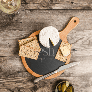 A round wooden cheese board with crackers, brie, sliced cheese, and a knife rests on a rustic table. A glass of white wine, pickles, and the White 5x7 Frame Mockup with Linen | Real Photo PSD complete the inviting display.
