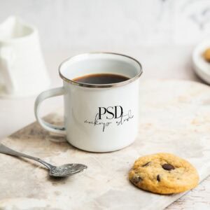 The Enamel Camp Mug Mockup | Cozy Kitchen Lifestyle PSD features a white enamel mug filled with coffee on a light marble surface, accompanied by a chocolate chip cookie and spoon, with cookies and milk softly blurred in the background.