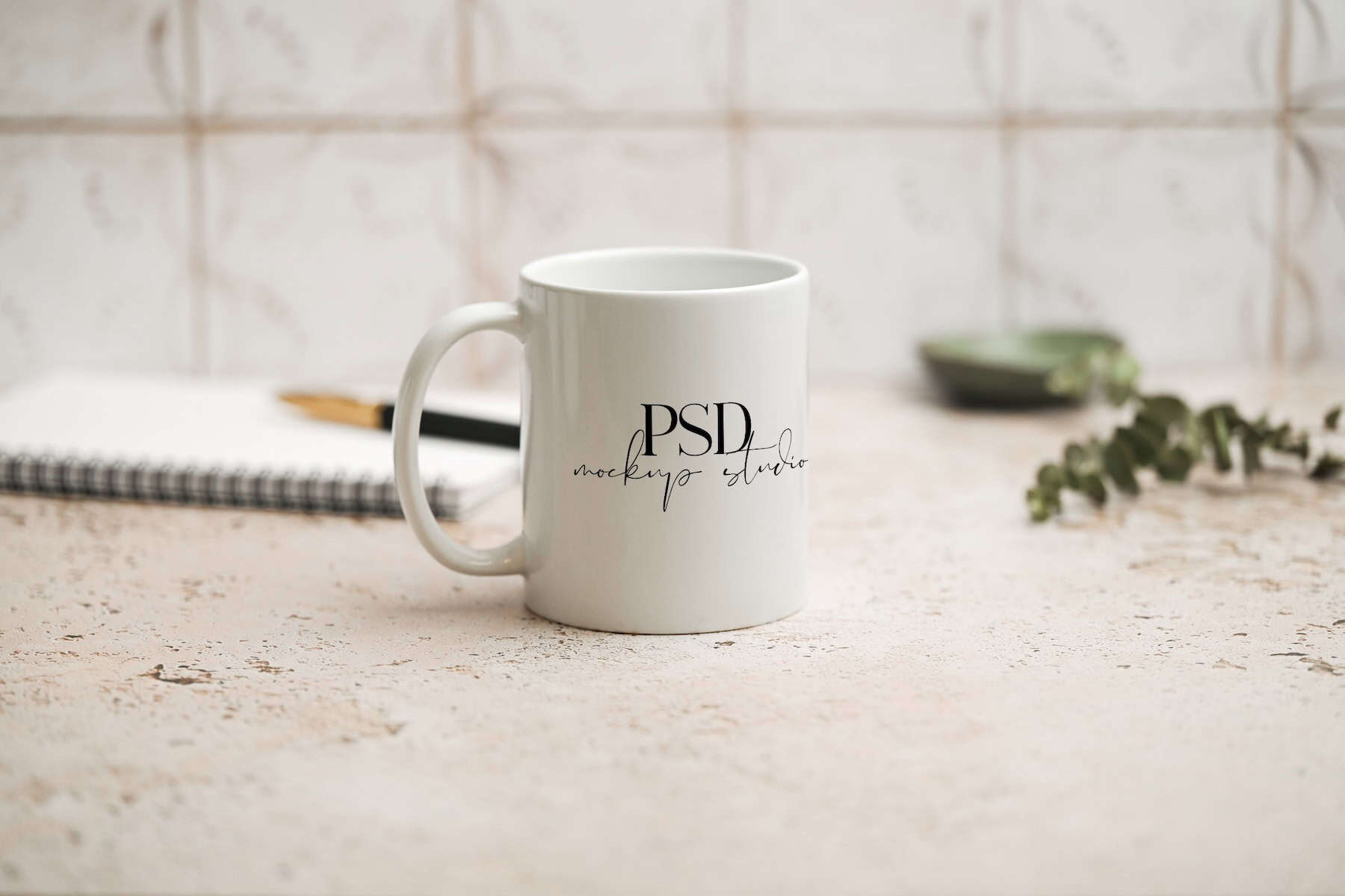A white ceramic mug with PSD mockup studio printed on it sits on a light-colored, textured surface near a notebook, pen, green bowl, and some greenery, with a patterned background.