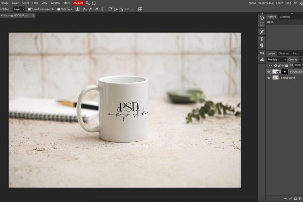 A Photoshop window displays a mockup of a white mug with PSD mockup studio text, placed on a textured surface with a notebook, pen, green plate, and leaves in the blurred background.