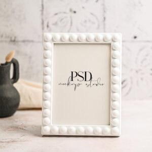White 5x7 Frame Mockup with Linen | Real Photo PSD displayed upright on a light surface, accented with raised circles. A dark vase and beige cloth are blurred in the background. The frame shows placeholder text reading PSD mockup studio.