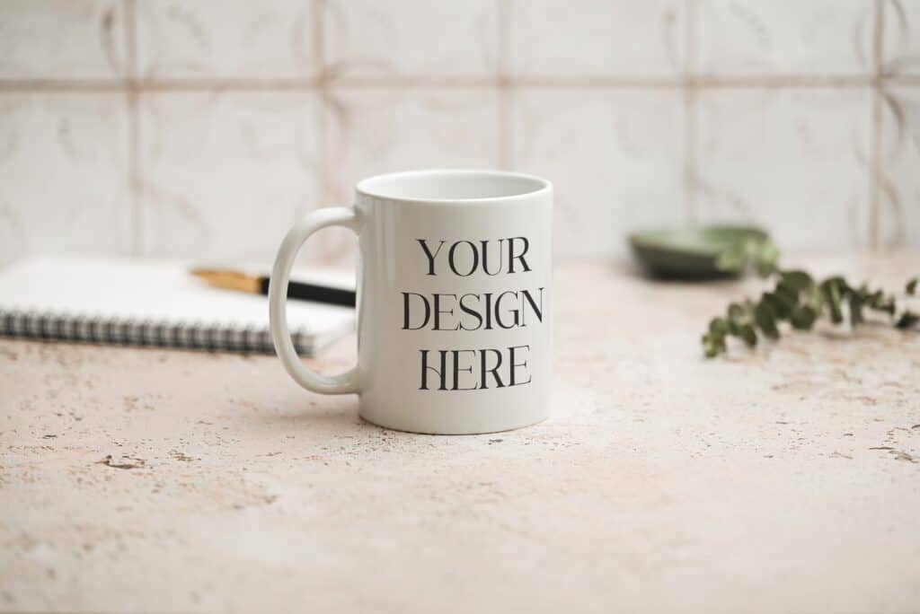 A white mug with the words YOUR DESIGN HERE printed on it sits on a light textured surface. In the background, there is a notebook, a pen, and some greenery, with a blurred, patterned wall behind them.