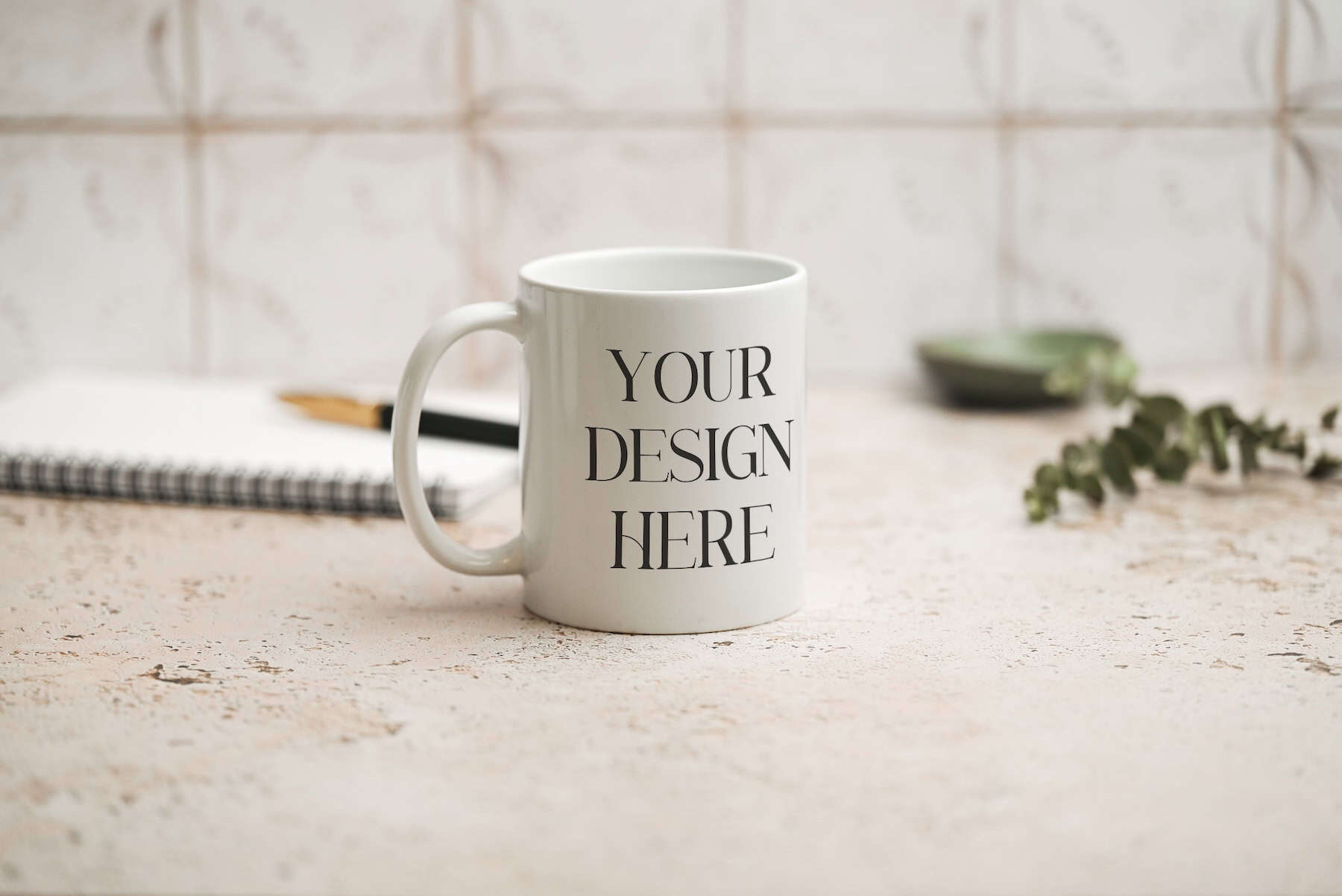 A white mug with the words YOUR DESIGN HERE printed on it sits on a light textured surface. In the background, there is a notebook, a pen, and some greenery, with a blurred, patterned wall behind them.