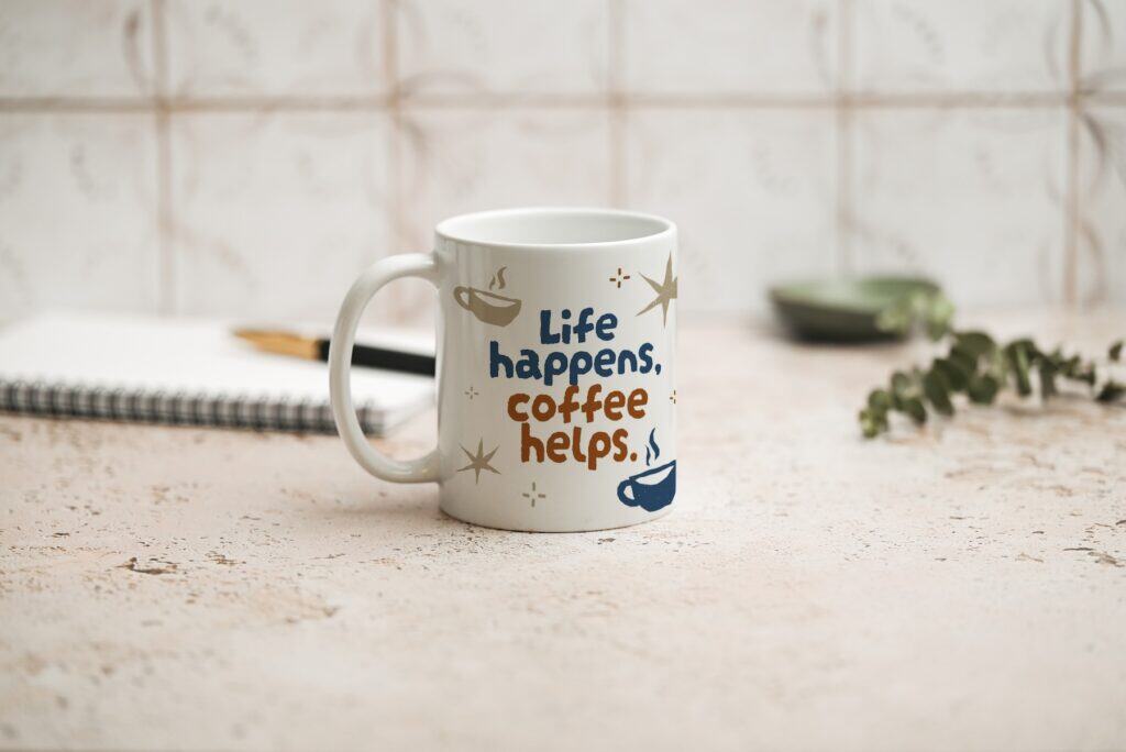 A white coffee mug with the text Life happens, coffee helps. sits on a light-colored table. In the background, there is a spiral notebook, a pen, a small green dish, and a sprig of greenery.