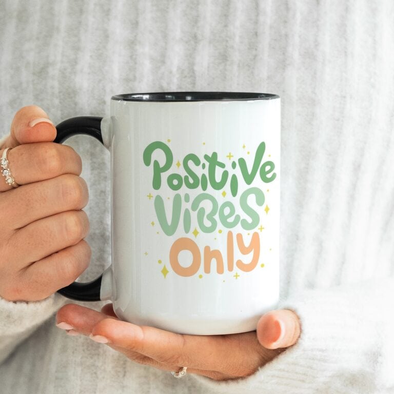 A person in a white sweater holds a Free 15oz Black Accent Mug Mockup featuring colorful "Positive Vibes Only" text in green and orange, with small yellow sparkles surrounding the words.