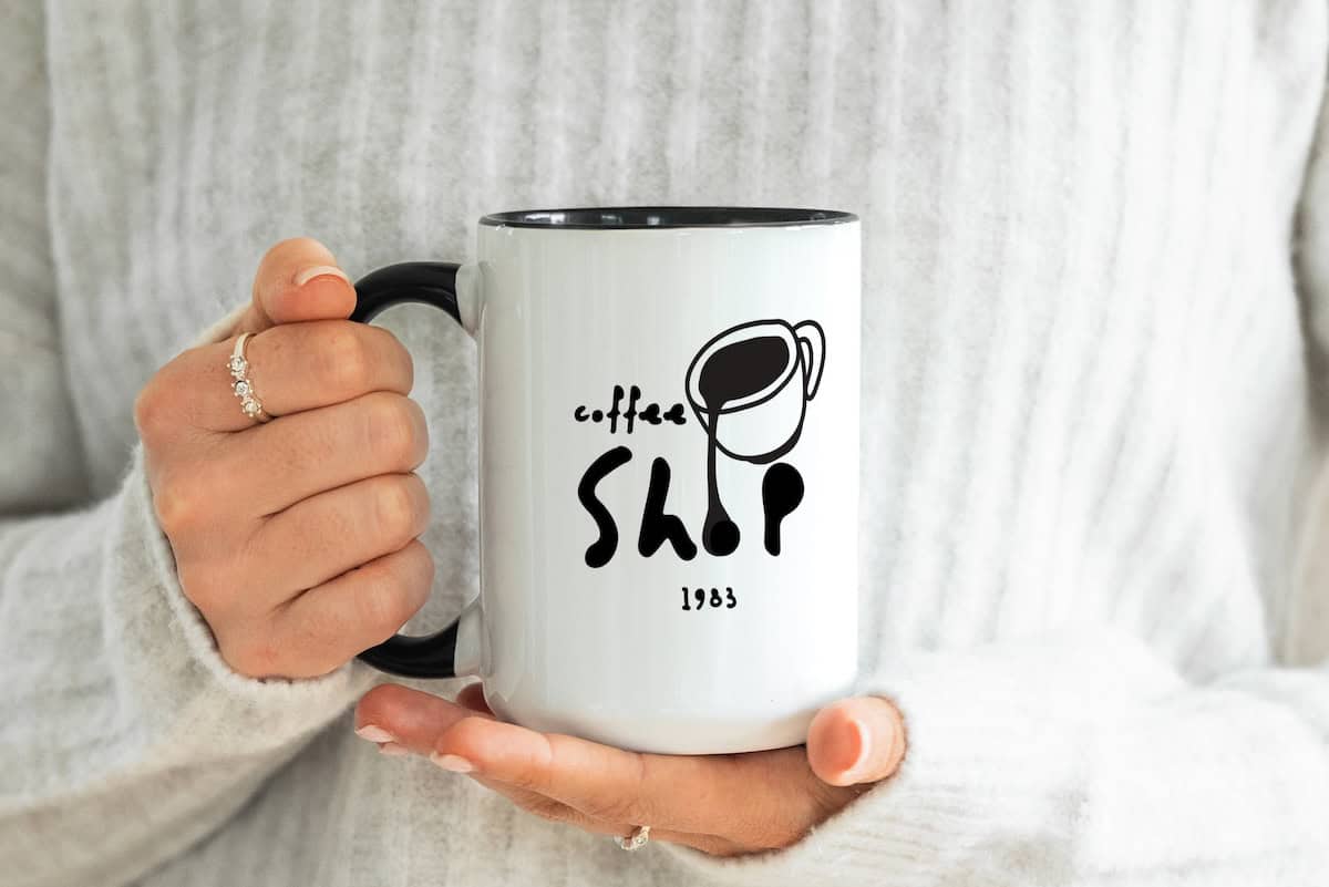 A person in a white sweater holding a white mug with black handle. The mug has a coffee cup graphic and the text coffee SHOP 1983 printed in black. The person has a ring on their finger.