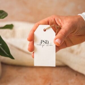 A hand holds a blank white tag with a twine loop. The tag has PSD mockup studio printed on it. A green plant and a beige fabric are blurred in the background.