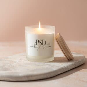 The Frosted Candle Mockup PSD 11oz | Real Photo shows a lit white candle in a frosted glass jar labeled PSD mockup studio on a stone tray with its wooden lid beside it, set against a soft beige background for an elegant look.