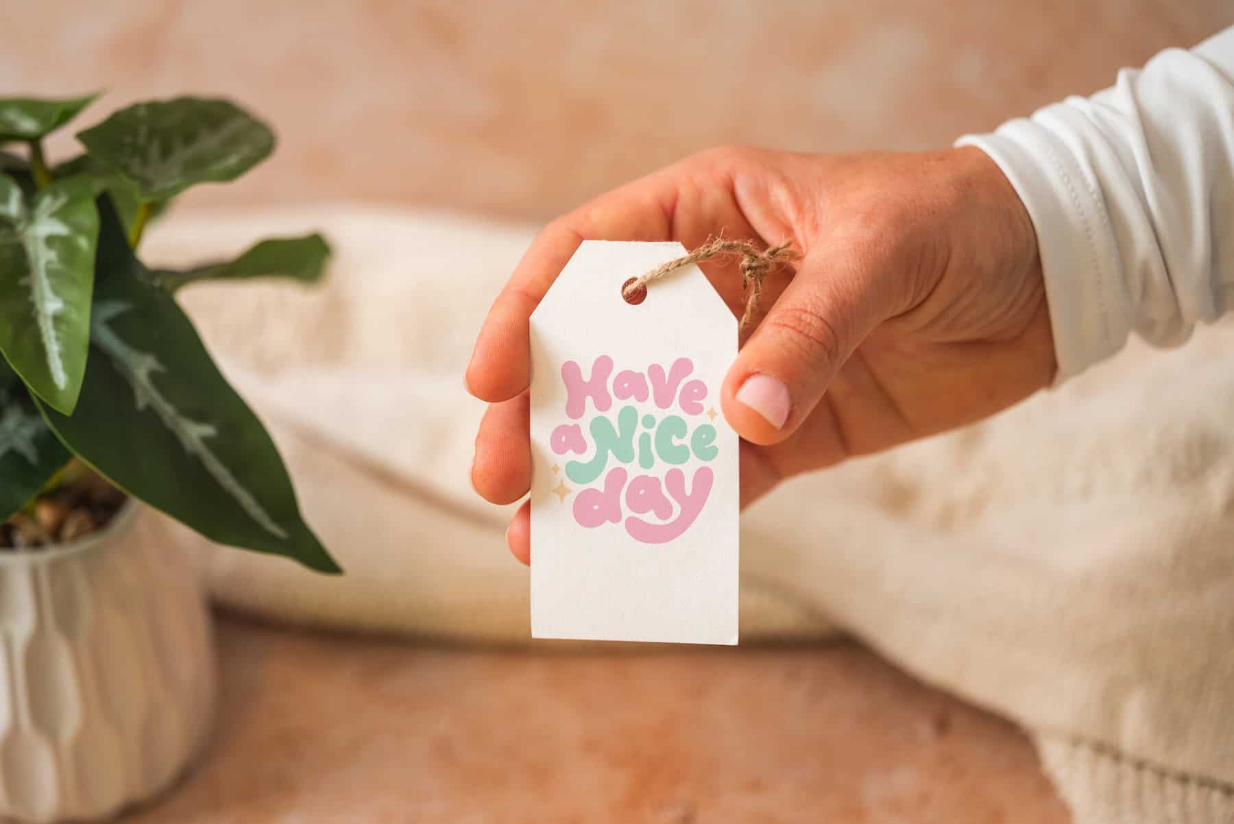 A hand holds a white tag with colorful text reading Have a nice day. A green potted plant and a beige fabric are in the background.