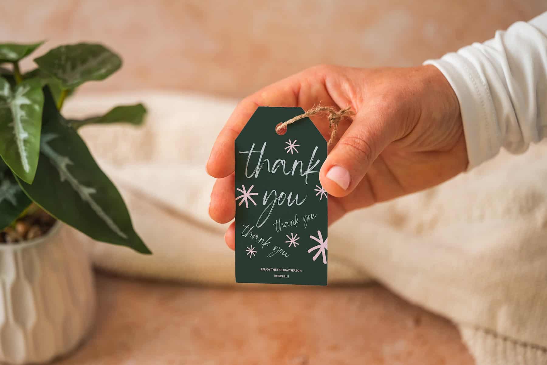 A hand holds a dark green gift tag tied with twine, featuring the words thank you written multiple times in white cursive and surrounded by simple flower illustrations. A potted plant is visible in the background.