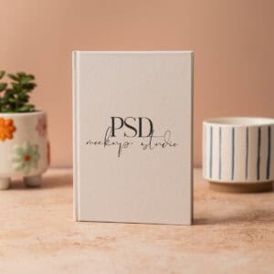 A 5x7 Hardcover Journal Mockup PSD stands upright on a beige surface, with two ceramic plant pots—one with flowers and one with blue stripes—in the background, all set against a soft brown wall in a warm neutral lifestyle scene.