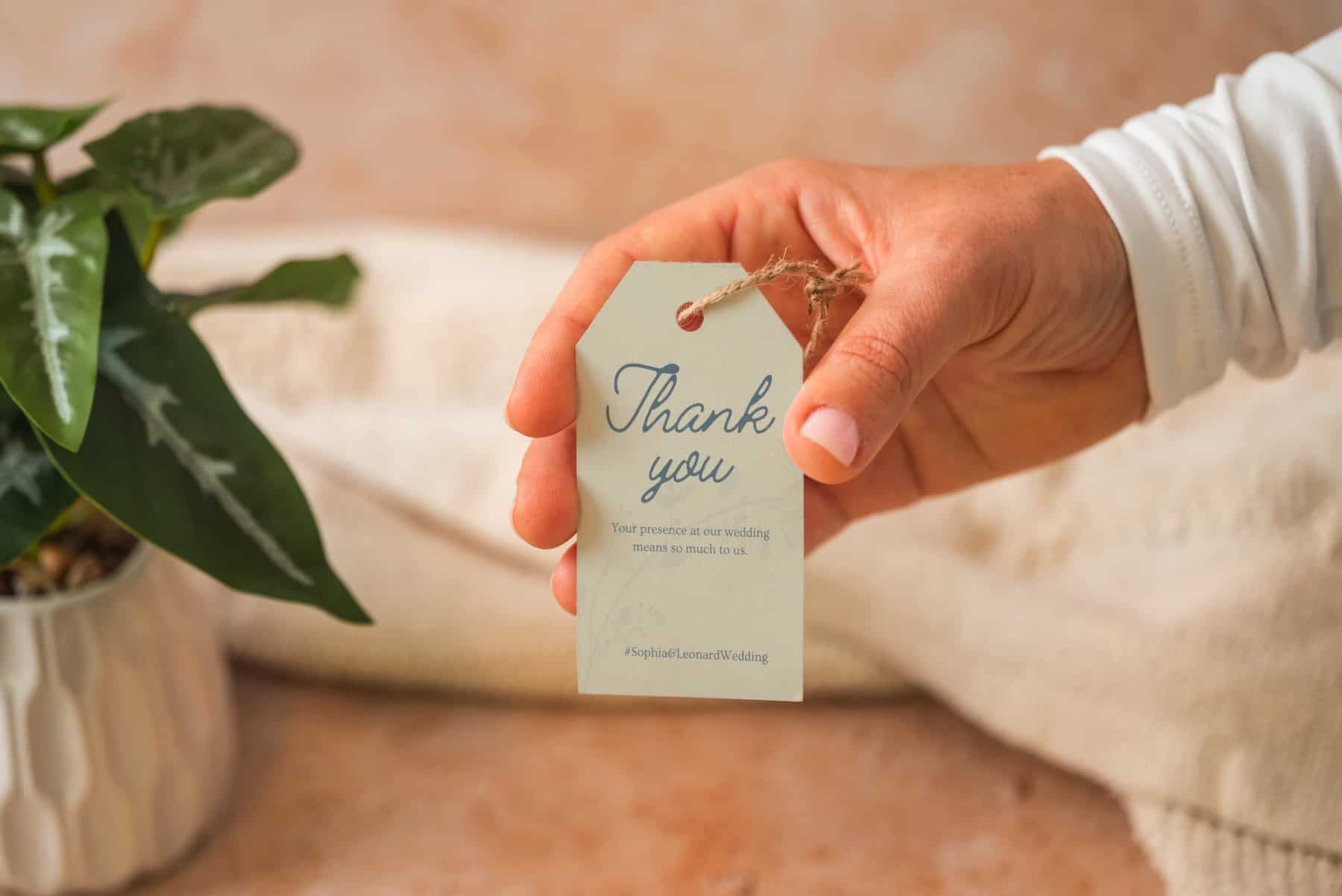 A hand holds a tag that reads Thank you. Your presence at our wedding means so much to us. Next to the hand is a potted green plant on a soft, beige surface.