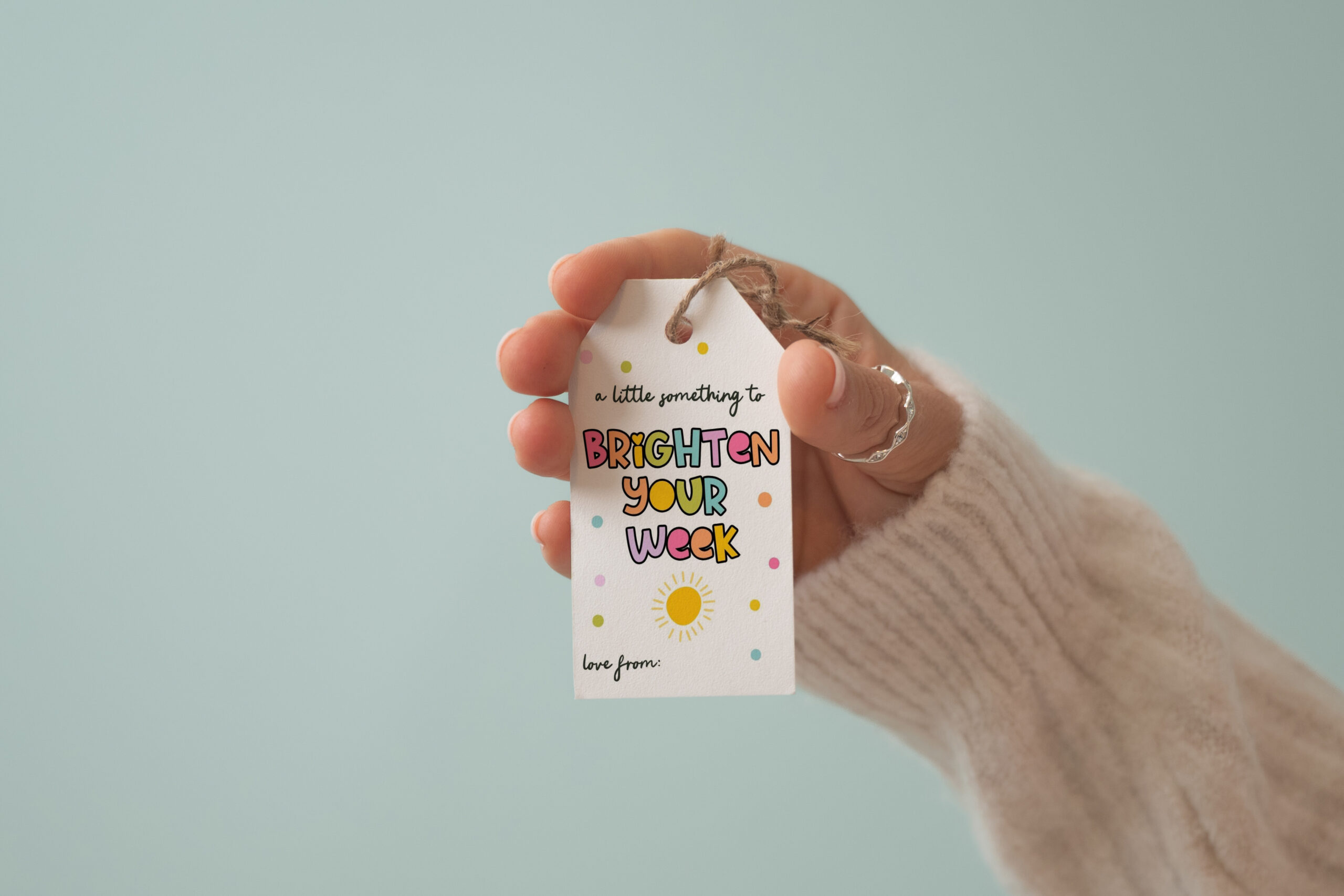 A hand in a cozy sweater holds a gift tag reading, A little something to BRIGHTEN YOUR WEEK, surrounded by colorful dots and a sun illustration, with love from: at the bottom. Perfect for showcasing your own designs, this Free Gift Tag Mockup PSD lets you easily personalize your thoughtful presents.