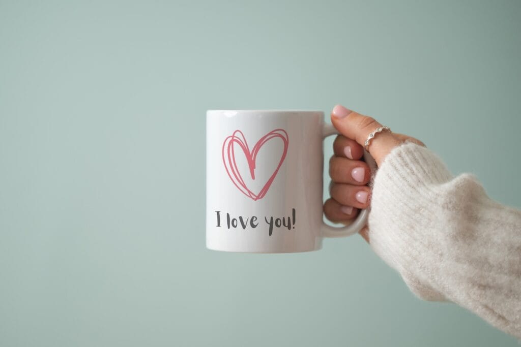 A hand in a cozy cream sweater holds a white mug featuring a pink heart illustration and the words I love you! against a soft light green background.