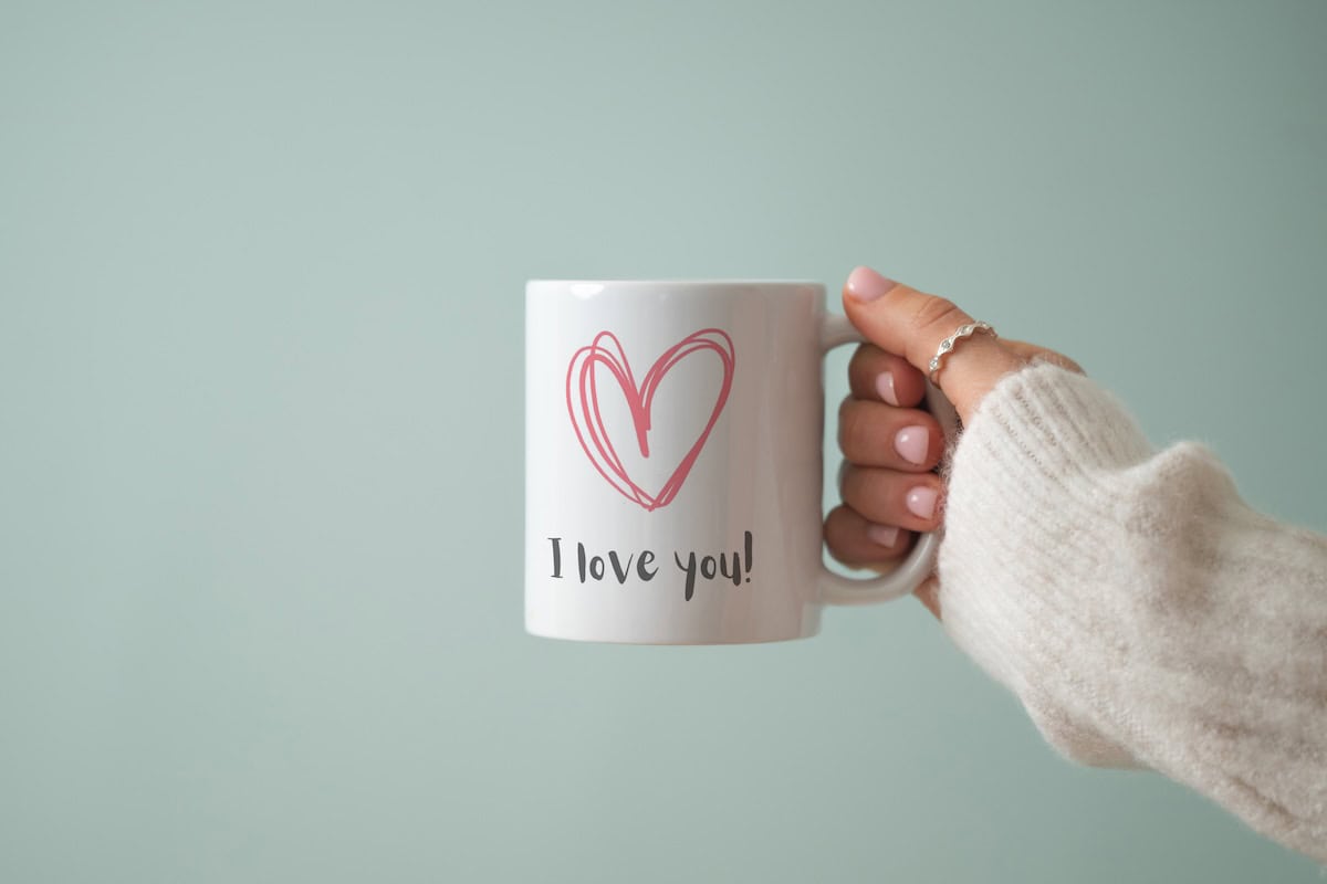 A hand in a cozy cream sweater holds a white mug featuring a pink heart illustration and the words I love you! against a soft light green background.