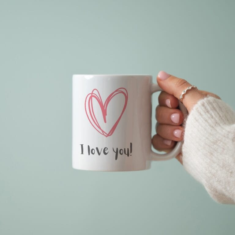 A hand wearing a white sweater holds a white mug with a pink heart and the words I love you!—the perfect Free 11oz Mug Mockup PSD—set against a light blue background.