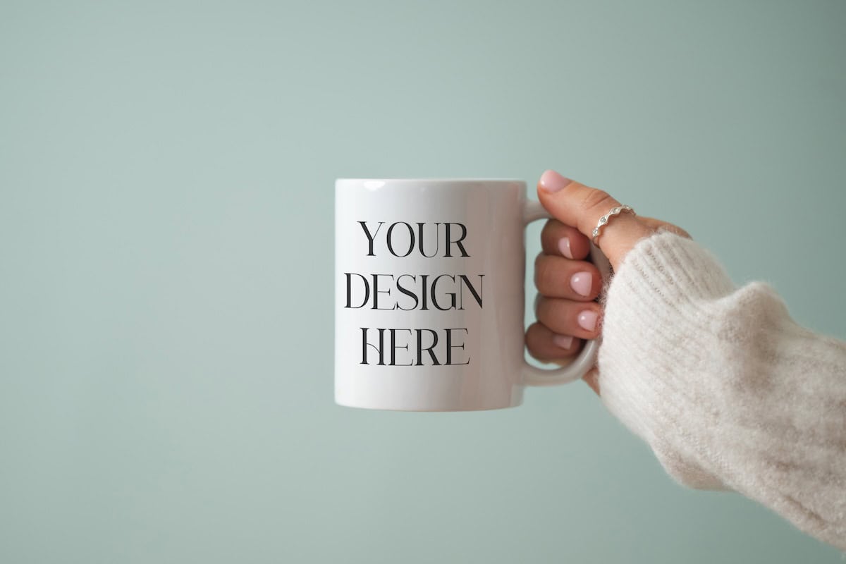 A hand in a cream-colored sweater holds a white mug against a light green background. The mug has the text YOUR DESIGN HERE printed in black, suggesting it’s customizable.