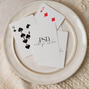 A white plate holds four cards—9 of spades, 5 of diamonds, and two face-down with "PSD mockup studio" text—alongside a candle on textured beige cloth. Shown: Poker Playing Card Mockup PSD | Real Photo Flat Lay.