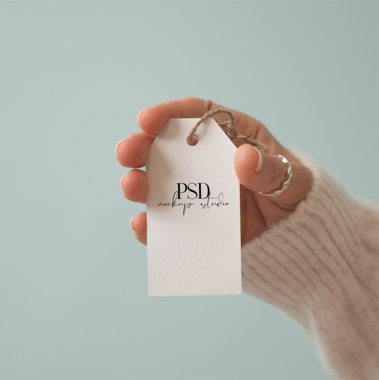 A hand wearing a cream sweater and a ring holds a blank white tag with PSD mockup studio written on it, against a plain light blue background—perfect for showcasing your designs with this Free Gift Tag Mockup PSD.