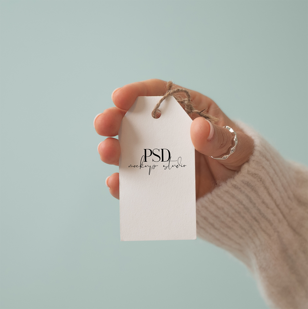 A hand wearing a cream sweater and a ring holds a blank white tag with PSD mockup studio written on it, against a plain light blue background—perfect for showcasing your designs with this Free Gift Tag Mockup PSD.