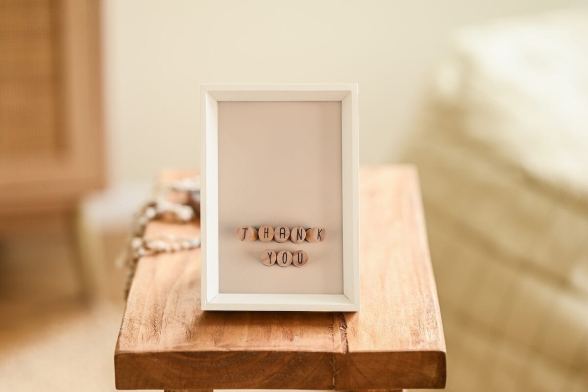 A white-framed picture with letter beads spelling THANK YOU sits on a wooden table in a softly lit, cozy room.