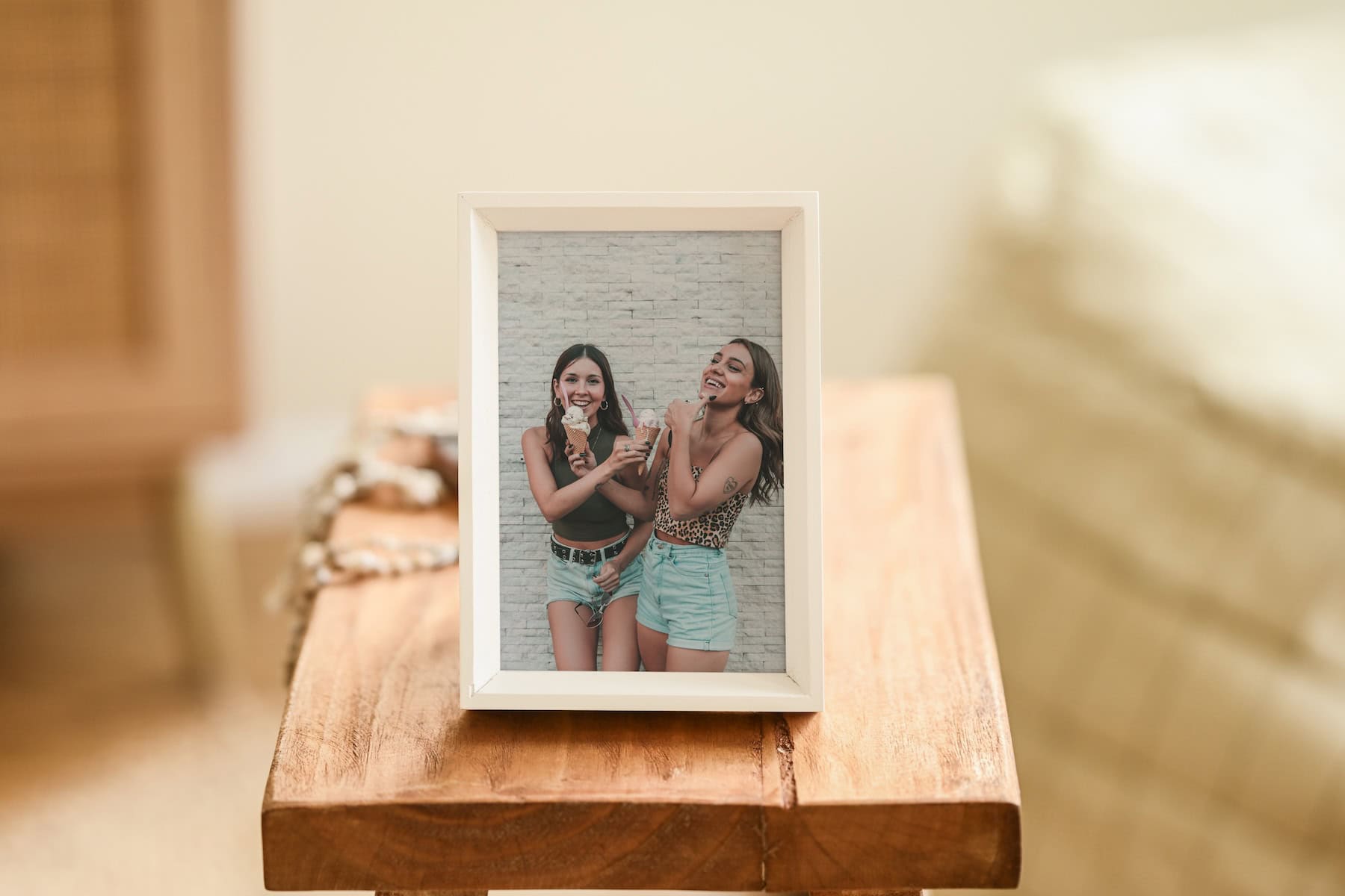 A framed photo of two young women laughing and standing close together, placed on a wooden table. The background is softly blurred, highlighting the framed picture as the focal point.