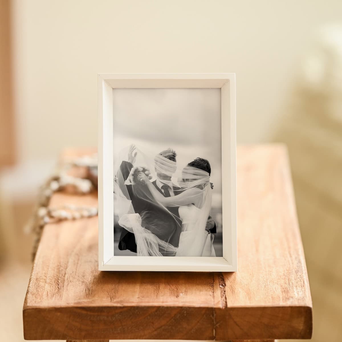 A framed black-and-white photo of a newlywed couple embracing, with the brides veil blowing around them, sits on a wooden table. The background is softly blurred.