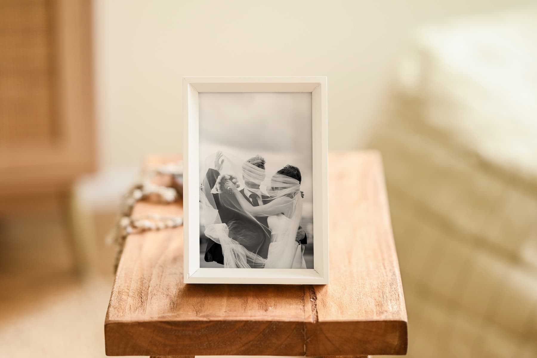 A framed black-and-white photo of a couple on their wedding day sits on a wooden table. The couple is embracing and smiling, partially covered by a veil, with soft, cozy surroundings in the background.