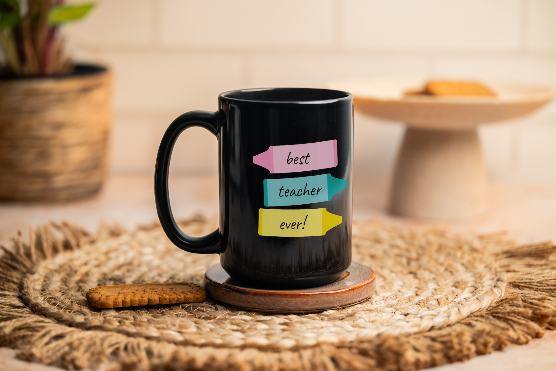 A black mug with colorful labels reading best teacher ever! sits on a woven mat. A biscuit rests beside the mug, with a plant and plate of cookies blurred in the background.