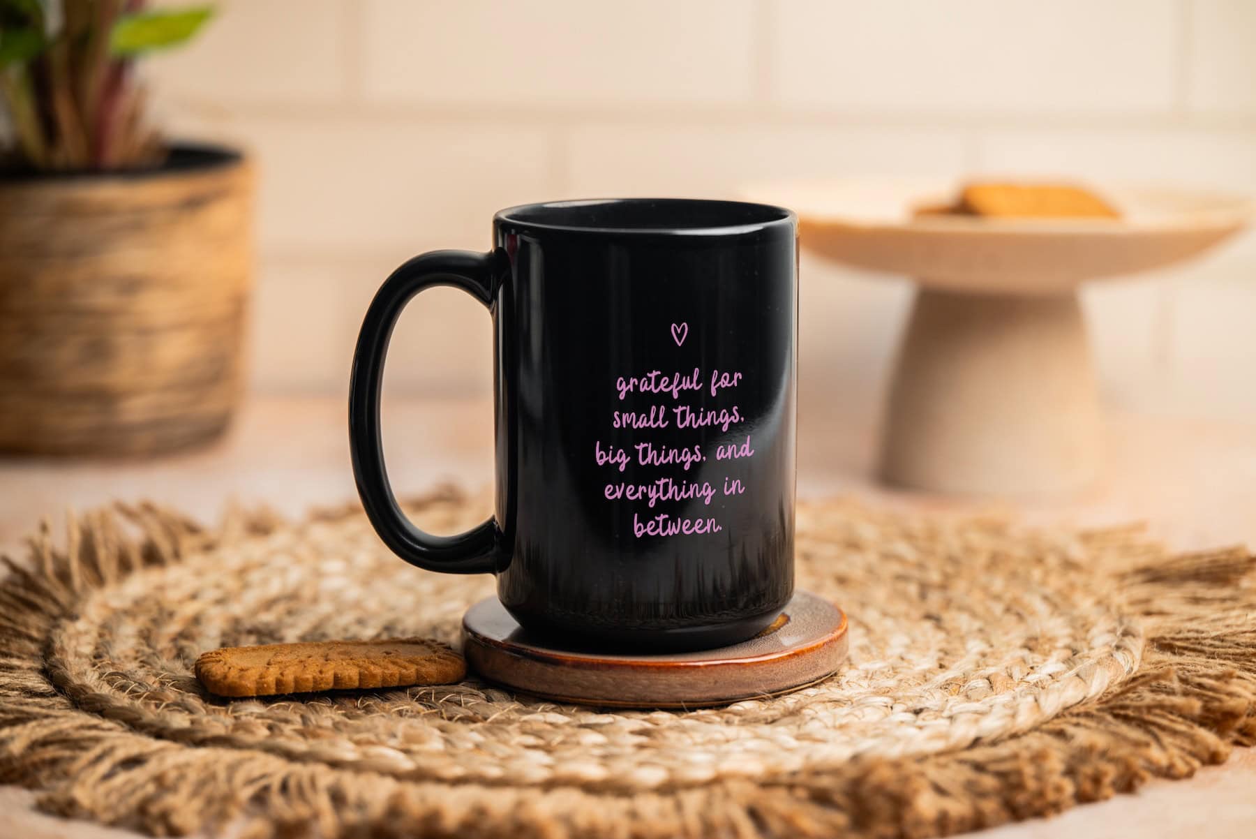 A black mug with pink text that reads, grateful for small things, big things, and everything in between, sits on a coaster atop a woven placemat. A biscuit lies beside it, with another plate and plant blurred in the background.